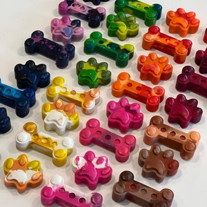 May include: Assortment of colorful crayon melts shaped like dog bones and paw prints. The crayons are arranged in a row on a white surface.