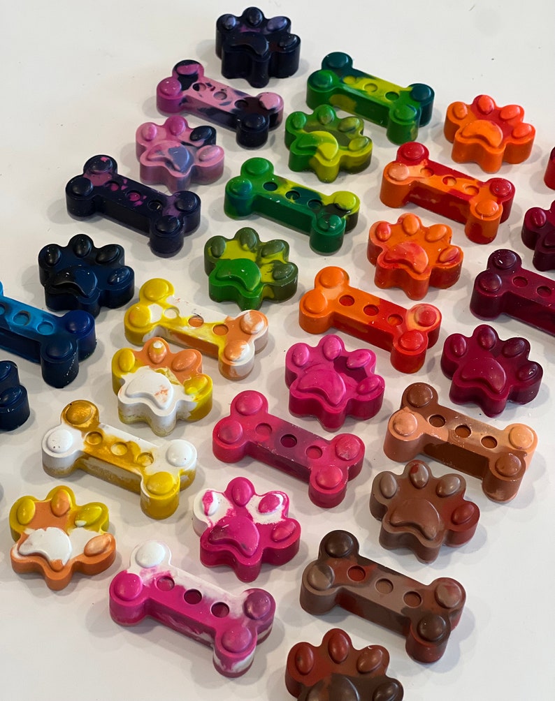 May include: Assortment of colorful crayon melts shaped like dog bones and paw prints. The crayons are arranged in a row on a white surface.