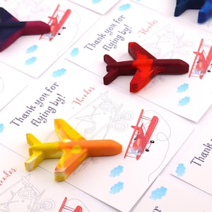 Puede incluir: Crayones con forma de avi&oacute;n en colores rojo, naranja, amarillo y azul, sobre tarjetas blancas. Las tarjetas presentan una ilustraci&oacute;n de biplano y el texto "Thank you for flying by!" con un nombre.