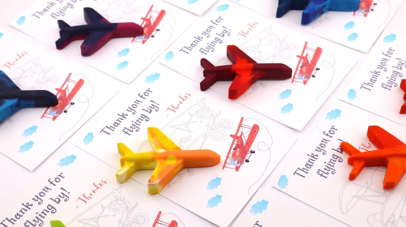 Puede incluir: Crayones con forma de avi&oacute;n en colores rojo, naranja, amarillo y azul, sobre tarjetas blancas. Las tarjetas presentan una ilustraci&oacute;n de biplano y el texto "Thank you for flying by!" con un nombre.