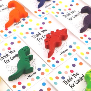 May include: A collection of colorful dinosaur-shaped crayons atop white cards. Each card has the text "Thank You For Coming!" and a name. The cards are decorated with colorful polka dots, creating a playful and festive look.