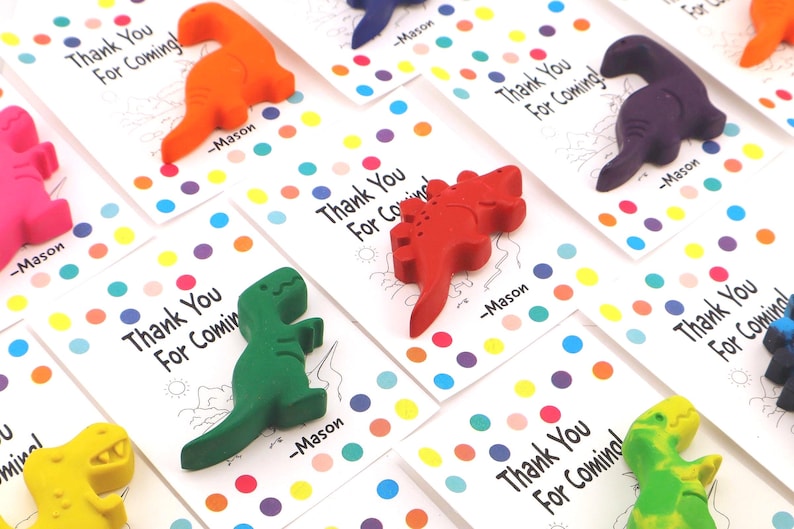 May include: A collection of colorful dinosaur-shaped crayons atop white cards. Each card has the text "Thank You For Coming!" and a name. The cards are decorated with colorful polka dots, creating a playful and festive look.