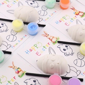 May include: An Easter-themed craft kit featuring white egg-shaped ornaments, paint pots in various pastel colors, paintbrushes, and coloring pages with the text "Happy Easter" and illustrations of bunnies and chicks. The kit is designed for creative activities.
