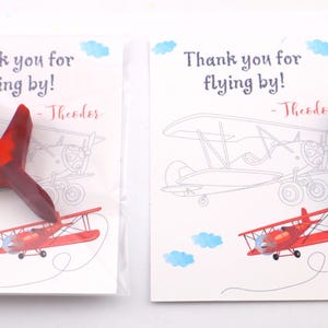 Puede incluir: Dos crayones con forma de avi&oacute;n, uno rojo y otro azul, descansan sobre tarjetas con ilustraciones de aviones y el texto "Thank you for flying by!" y un nombre. Las tarjetas tienen acentos de nubes azules.