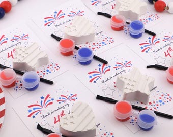 May include: A collection of craft kits for the 4th of July. Each kit includes a white star or heart-shaped plaster piece, red and blue paint, a paintbrush, and a card with patriotic graphics and text.