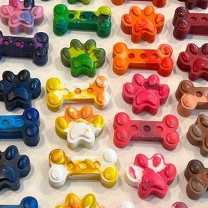May include: Assortment of colorful dog bone and paw-shaped crayons. The crayons are made in a variety of colors, including red, orange, yellow, green, blue, and purple. The crayons are arranged in a grid pattern on a white surface.