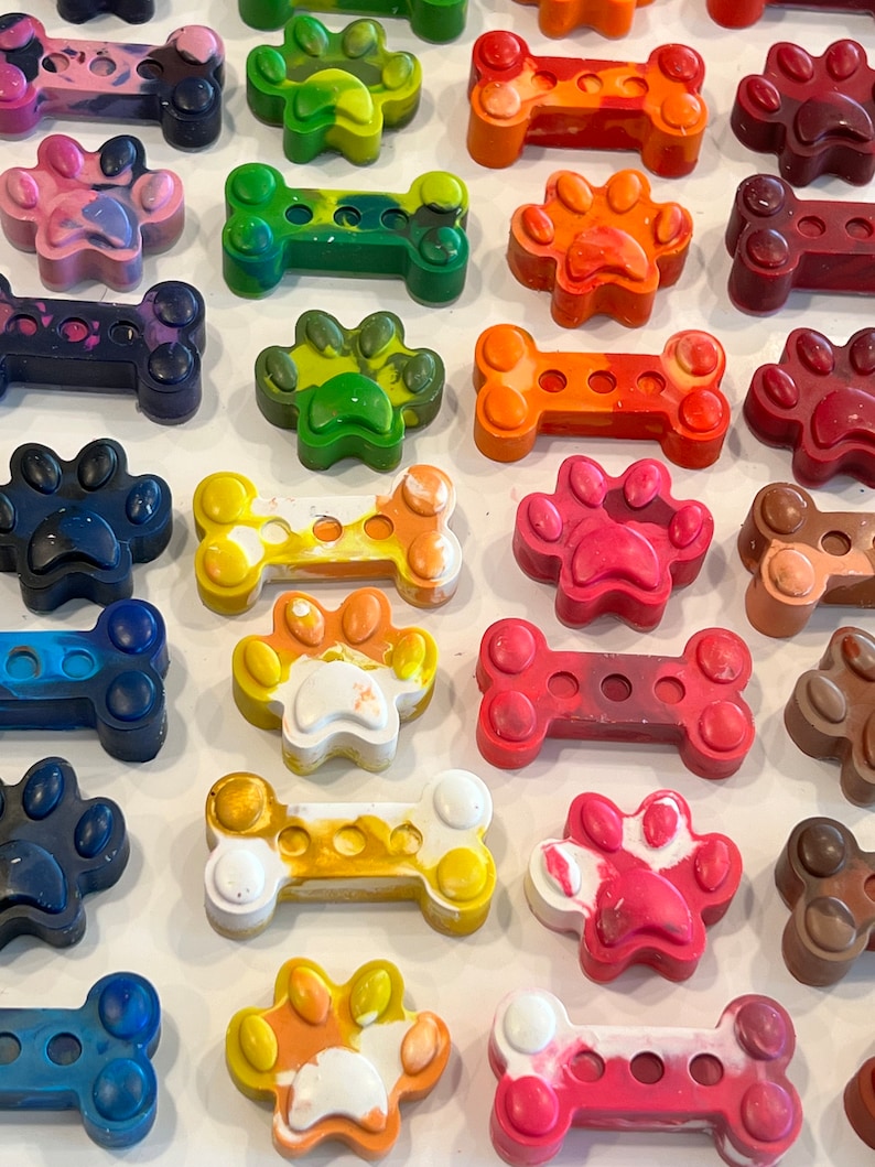 May include: Assortment of colorful dog bone and paw-shaped crayons. The crayons are made in a variety of colors, including red, orange, yellow, green, blue, and purple. The crayons are arranged in a grid pattern on a white surface.