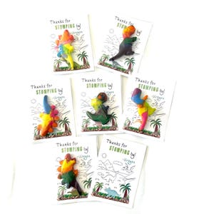 May include: A collection of handmade dinosaur-shaped crayons in various colors, displayed on white cards. Each card features the text "Thanks for STOMPING by!" and a small illustration of a dinosaur scene. The crayons are a mix of vibrant hues.