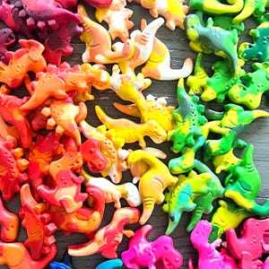 May include: A collection of colorful dinosaur-shaped crayons in a rainbow of hues, including red, orange, yellow, green, blue, and pink. The crayons are arranged on a wooden surface, creating a vibrant and playful display.
