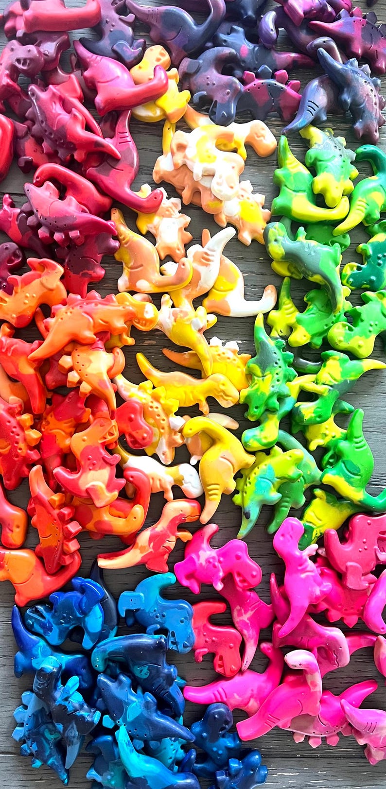 May include: A collection of colorful dinosaur-shaped crayons in a rainbow of hues, including red, orange, yellow, green, blue, and pink. The crayons are arranged on a wooden surface, creating a vibrant and playful display.
