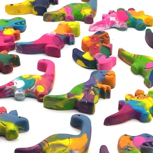 May include: A collection of colorful dinosaur-shaped crayons in various shades of pink, blue, yellow, green, and orange. The crayons are arranged on a white surface, showcasing their vibrant, multi-colored appearance. These crayons are ideal for children's art projects.