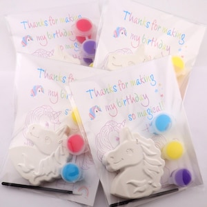 May include: Unicorn-shaped craft kits for a birthday party. Each kit includes a white unicorn figure, paint pots in various colors, and a paintbrush. The packaging reads "Thanks for making my birthday so magical!"