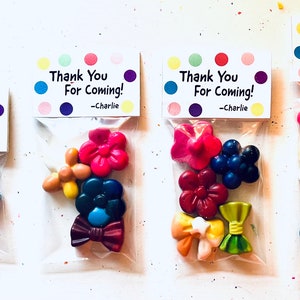 Puede incluir: Seis pequeñas bolsas de crayones de cera de colores con forma de flores y lazos. Cada bolsa tiene una etiqueta de agradecimiento con el texto "¡Gracias por venir!" y un nombre en la parte inferior.