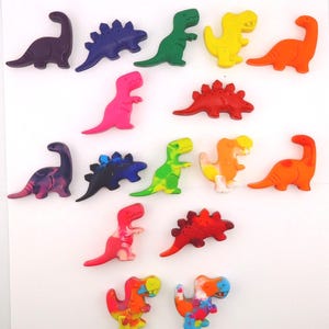 May include: A collection of colorful dinosaur-shaped crayons in various shades, including purple, blue, green, yellow, orange, and pink. The crayons are arranged on a white surface, showcasing their different shapes and sizes. These crayons are perfect for creative projects.