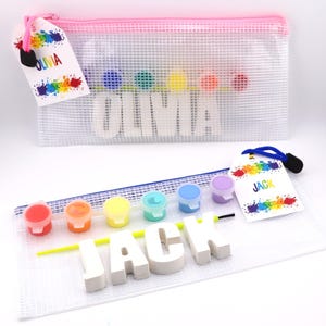May include: Two clear mesh pencil cases with pink zippers. One case has the name "OLIVIA" and the other has "JACK" in white block letters. Each case contains paint pots and a paint brush. Colorful paint splatters decorate the tags.