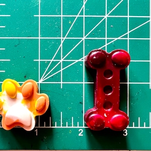 May include: Two crayon shapes on a green cutting mat. One is a paw print in yellow, orange, and white. The other is a bone shape in red.