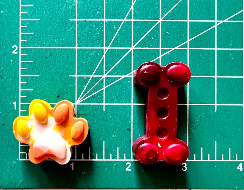 May include: Two crayon shapes on a green cutting mat. One is a paw print in yellow, orange, and white. The other is a bone shape in red.