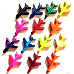 Puede incluir: Una colecci&oacute;n de ceras con forma de avi&oacute;n de colores vibrantes, incluyendo morado, azul, verde, amarillo, rosa, rojo y naranja. Las ceras est&aacute;n dispuestas sobre una superficie blanca, mostrando sus colores vibrantes y su dise&ntilde;o l&uacute;dico.