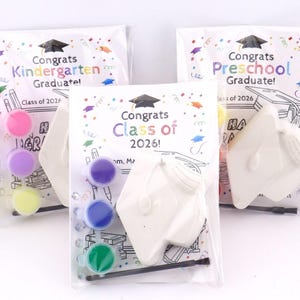 May include: Three graduation-themed craft kits in clear plastic bags. Each kit includes a ceramic mortarboard, paint pots in pink, purple, yellow, blue, and green, and a paintbrush. The bags are labeled "Congrats Kindergarten Graduate!", "Congrats Class of 2026!", and "Congrats Preschool Graduate!"