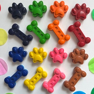 May include: Assortment of colorful crayons shaped like dog paws and bones. The crayons are arranged in two rows on a white surface.