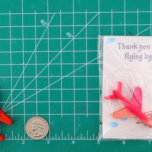 Puede incluir: Una esterilla de corte verde muestra un avi&oacute;n de juguete rojo, una moneda y una tarjeta. La tarjeta dice "Thank you for flying by!" con una ilustraci&oacute;n de avi&oacute;n. El avi&oacute;n es rojo con alas rojo oscuro. Medidas en cent&iacute;metros.