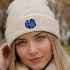Könnte beinhalten: Eine cremefarbene Strickmütze mit einem kleinen, gestickten Pixel-Art-Design eines blauen Huhns auf dem vorderen Umschlag. Die Mütze besteht aus weichem Rippstrickmaterial und hat einen umgeschlagenen Rand, der fest am Kopf anliegt.