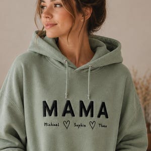 Könnte beinhalten: Ein salbeigrüner Kapuzenpullover mit dem Wort MAMA, das in fetten schwarzen Buchstaben auf die Brust gestickt ist. Unter dem Wort sind die Namen Michael, Sophie und Theo in Schwarz gestickt, getrennt durch zwei kleine schwarze Herzumrisse.