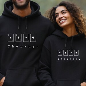 This black hoodie has a white image of playing cards centred in the middle, with the word therapy underneath. Tailored towards people and couples who enjoy playing cards games also who find playing card games a form of therapy.