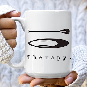 May include: White ceramic coffee mug with a black silhouette of a paddleboard and paddle. The text "Therapy." is printed below the image.