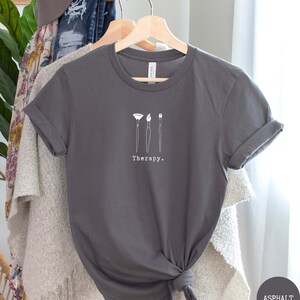 This asphalt t-shirt has a white image of three paintbrushes centred in the middle, with the word therapy underneath. Tailored towards people who enjoy painting also who find painting a form of therapy.