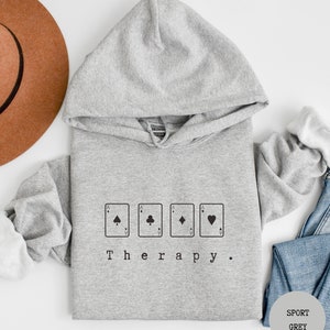 May include: A grey hooded sweatshirt with the text "Therapy." printed on the front. The text is surrounded by four playing card symbols: a spade, a club, a diamond, and a heart.