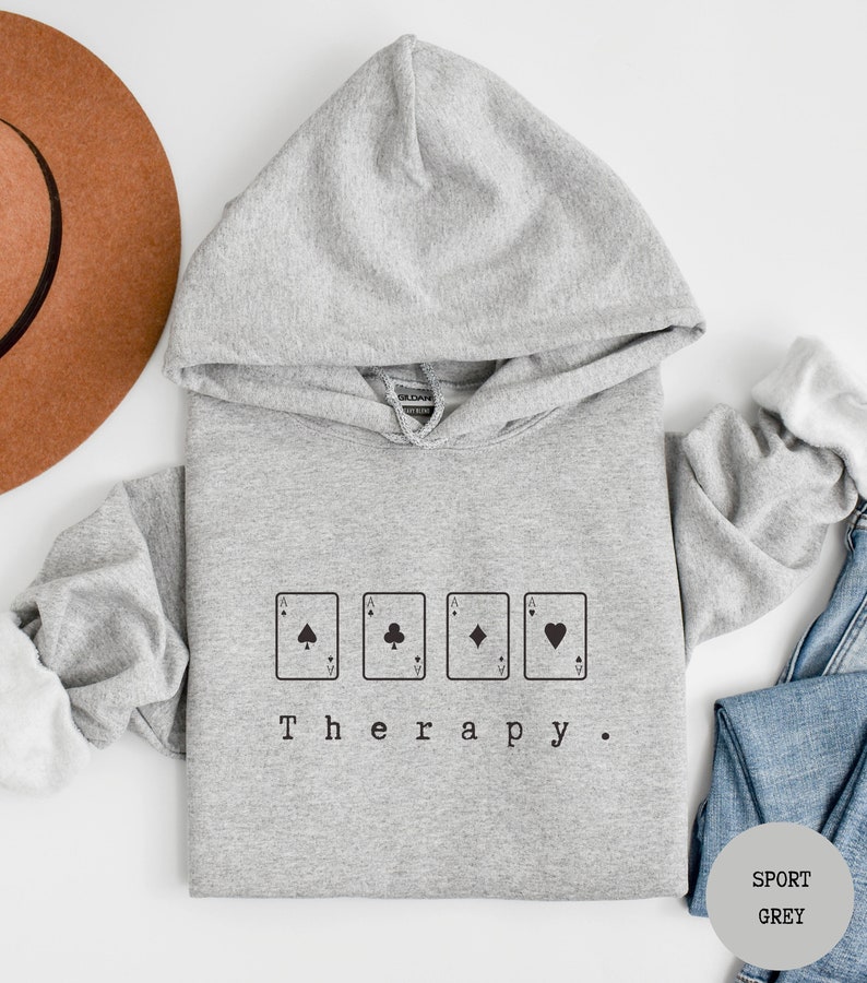 May include: A grey hooded sweatshirt with the text "Therapy." printed on the front. The text is surrounded by four playing card symbols: a spade, a club, a diamond, and a heart.