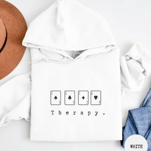 This white hoodie has a black image of playing cards centred in the middle, with the word therapy underneath. Tailored towards people and couples who enjoy playing cards games also who find playing card games a form of therapy.