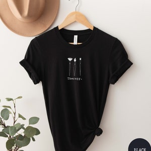 This black t-shirt has a white image of three paintbrushes centred in the middle, with the word therapy underneath. Tailored towards people who enjoy painting also who find painting a form of therapy.