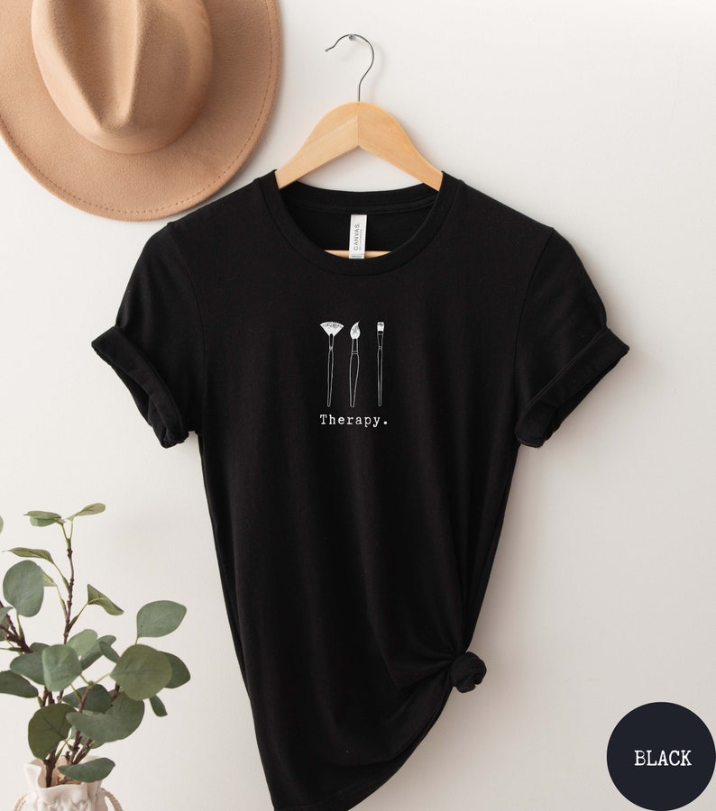 This black t-shirt has a white image of three paintbrushes centred in the middle, with the word therapy underneath. Tailored towards people who enjoy painting also who find painting a form of therapy.