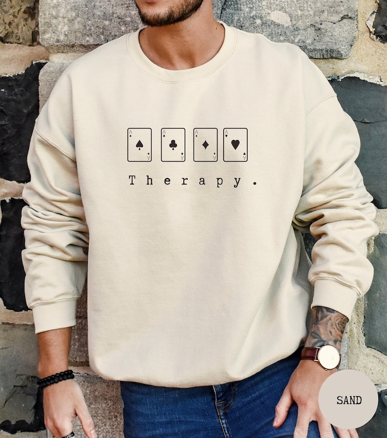 May include: A beige sweatshirt with a graphic of four playing cards, each showing an ace of a different suit. The text "Therapy." is printed below the cards.