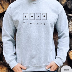 This sport grey sweatshirt has a black image of a playing cards centred in the middle, with the word therapy underneath. Tailored towards people who enjoy playing cards also who find card games a form of therapy