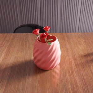 May include: A pink ceramic vase with a swirl design, holding three red paper roses.