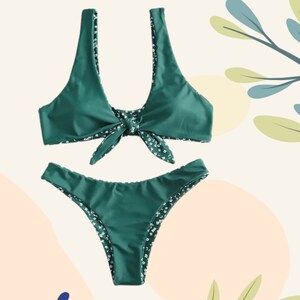 May include: A two-piece bikini set in a solid dark green color. The top features a front tie knot detail and wide shoulder straps. The bikini bottom has a high-cut leg design. Both pieces are lined with a contrasting white floral print fabric.