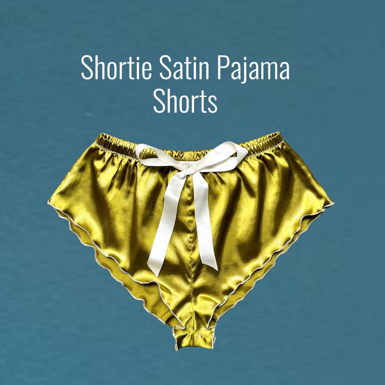 May include: Gold satin pajama shorts with a white satin bow. The shorts have a scalloped edge.