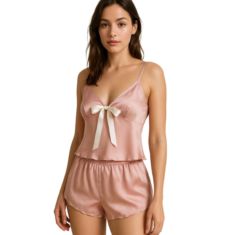May include: A blush pink satin pajama set with a bow detail on the top. The set includes a cami top and shorts.