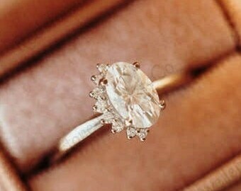May include: A close-up of a silver ring with an oval-shaped diamond in a halo setting. The ring is in a pink velvet ring box.