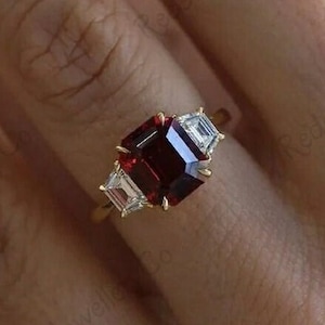 Puede incluir: Un anillo de oro con una piedra preciosa roja cuadrada y dos piedras blancas más pequeñas a cada lado.