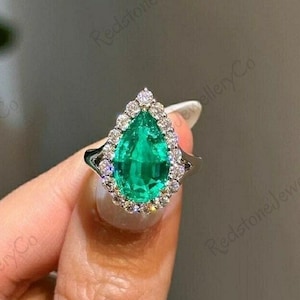 Può includere: Un anello di fidanzamento con smeraldo a forma di pera con un alone di diamanti più piccoli incastonati in una fascia d'argento.