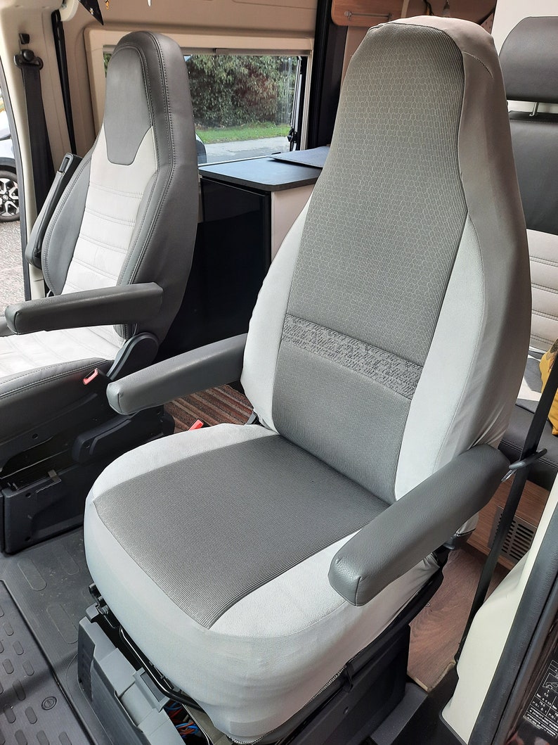 May include: A grey and white vehicle seat with armrests. The seat features a textured grey central panel and a lighter grey border. The armrests are a solid grey colour. The seat is in a vehicle interior.