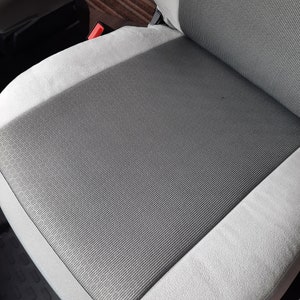 May include: A grey and white car seat with a textured, striped central panel. The seat has a light grey border and a red seatbelt buckle is visible. The seat appears to be clean and well-maintained.