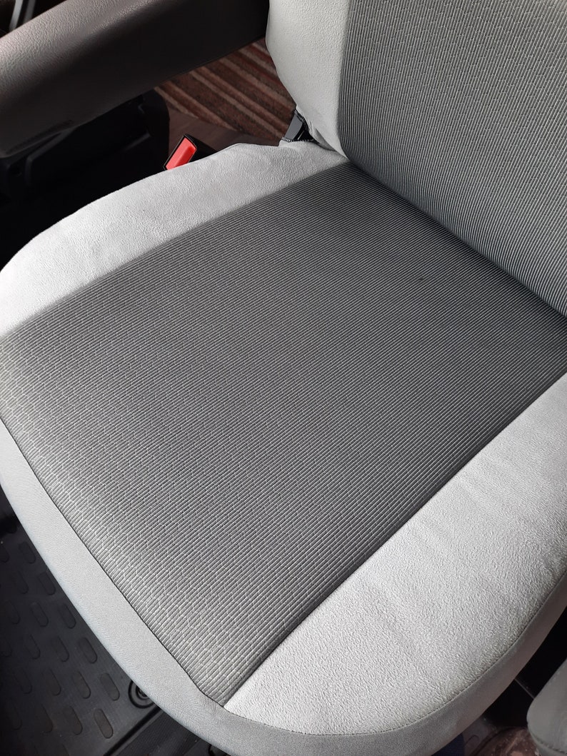 May include: A grey and white car seat with a textured, striped central panel. The seat has a light grey border and a red seatbelt buckle is visible. The seat appears to be clean and well-maintained.