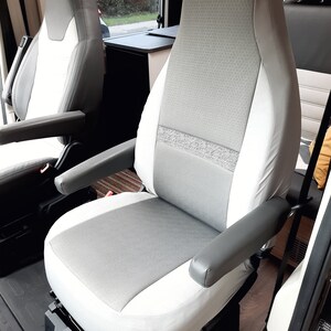 May include: A light grey and white vehicle seat with grey armrests. The seat features a textured grey central panel and a white border. The armrests are a solid grey colour. The seat is in a vehicle interior.