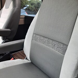 May include: Close-up of a grey and white vehicle seat. The seat has a textured grey fabric centre with a patterned band. The sides are a lighter grey. The seat is in a vehicle interior with a window visible in the background.