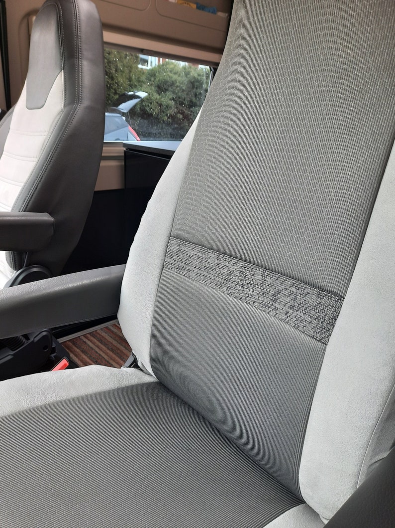 May include: Close-up of a grey and white vehicle seat. The seat has a textured grey fabric centre with a patterned band. The sides are a lighter grey. The seat is in a vehicle interior with a window visible in the background.